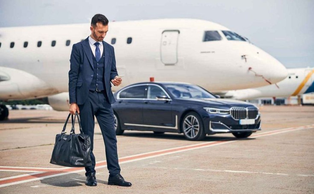 Corporate businessman holding a duffel bag and checking his phone next to a luxury sedan and private jet on the tarmac. Executive-Airport-Car-Service-Private-Jet